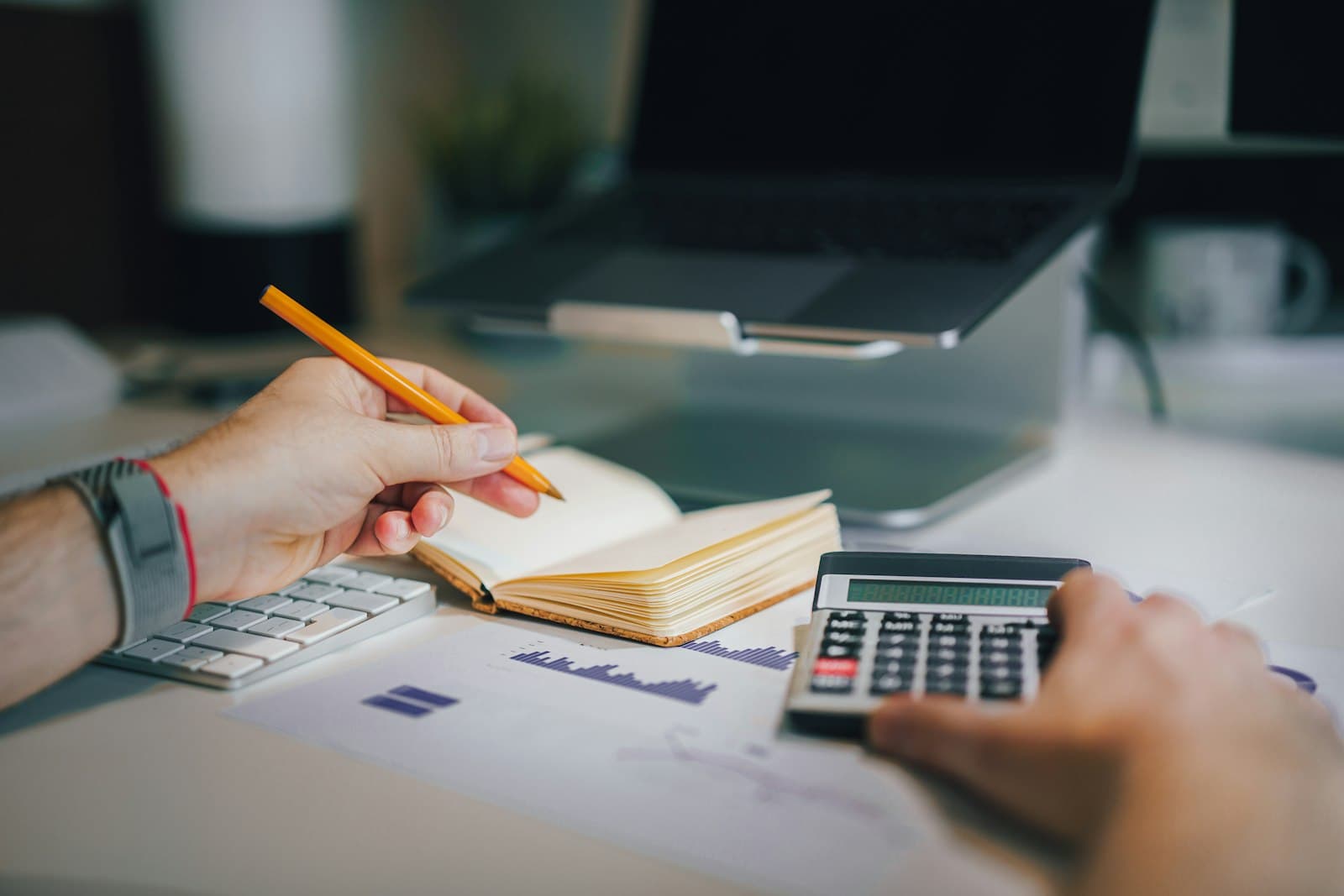 Hands recording figures in a notebook beside a calculator and financial charts